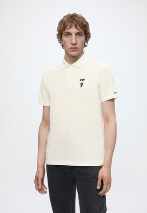 Joven con un polo de manga corta color crema con una pequeña figura de dibujo animado en blanco y negro en el pecho y pantalones oscuros.