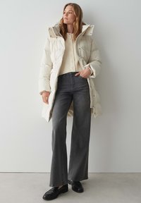 Manteau long et bouffant blanc avec capuche, pull crème à boutons et jean à pattes d'éléphant gris foncé. Des mocassins noirs complètent la tenue.