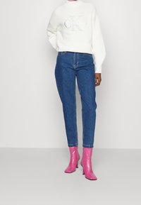 Pull blanc texturé avec le logo "CK", associé à un jean en denim bleu taille haute et des bottines à talons roses brillantes. Tenue simple et moderne.
