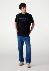 Black cotton T-shirt with "GUESS EST. 1981" logo; paired with blue denim jeans and light-colored sneakers; stands against a neutral background.