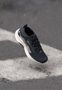 Svarte trening sko med teksturert overdel, grå mesh paneler, oransje detaljer og en hvit såle. Lett design med en strømlinjeformet fasong.