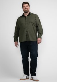 Camisa de botones verde estampada con mangas largas, combinada con jeans oscuros y zapatillas blancas con acentos negros.