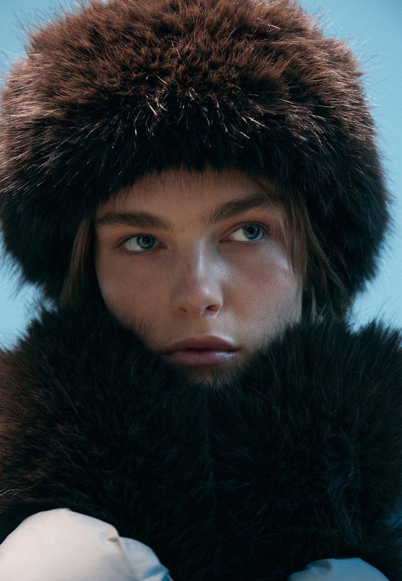 Cappello di pelliccia marrone con texture soffice, abbinato a una sciarpa di pelliccia coordinata. Pelle liscia e occhi azzurri visibili in primo piano. Illuminazione morbida.