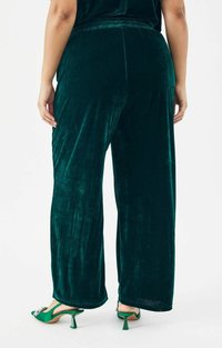 Flonkerige, wijde broek van fluweel in een rijke smaragdgroene kleur met een elastische tailleband. Soepele textuur, volledige lengte, en gecombineerd met groene schoenen.