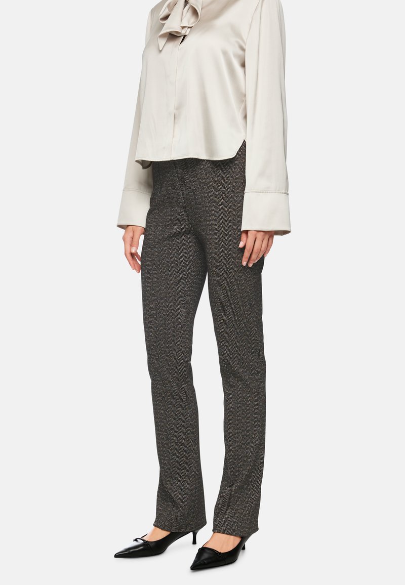 Eine Frau trägt hoch taillierte, dunkelgraue Fischgrät-Hosen, eine hellbeige Bluse mit Schleife und schwarze, spitze Kitten-Heels und steht vor einem weißen Hintergrund.