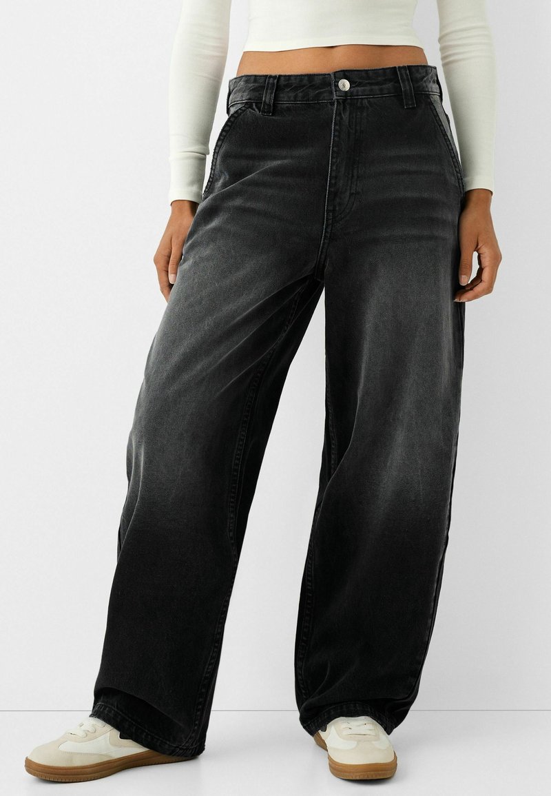 Personne portant un jean en denim noir ample et délavé, un crop top blanc à manches longues et des baskets blanches avec semelles en gomme, debout devant un fond uni.