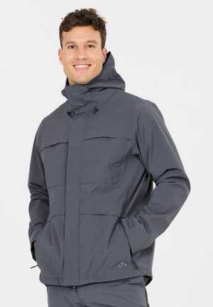 Mann lächelnd, trägt eine graue wasserdichte Kapuzenjacke mit vorderen Taschen und hat die Hände in den Seitentaschen, steht vor einem einfarbigen Hintergrund.