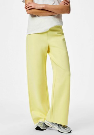 Osoba oblečená v svetlo žltých nohaviciach s širokými nohavicami, bielom tričku s krátkym rukávom a strieborno-šedých bežeckých topánkach stojaca so založenými ramenami.