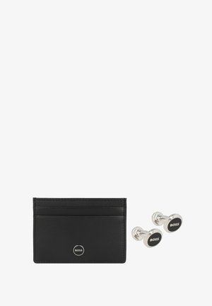 Tarjetero de cuero negro con múltiples ranuras y un juego de gemelos plateados circulares que presenta un logo negro en el centro.