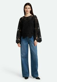 Blusa nera in pizzo con intricati motivi floreali e maniche a campana, abbinata a jeans in denim blu a vita alta. Texture liscia, vestibilità casual.