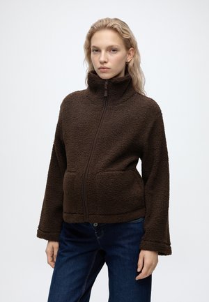 Jeune femme aux cheveux clairs porte une veste polaire marron foncé à fermeture éclair et un jean bleu, debout devant un fond clair uni.