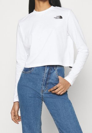 Femme portant un haut court blanc à manches longues avec le logo The North Face et un jean bleu taille haute, main reposant près de la poche.