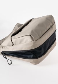 Beige schoudertas met een zwart gaas ritsvak aan de voorkant en een verstelbare band op een witte achtergrond.