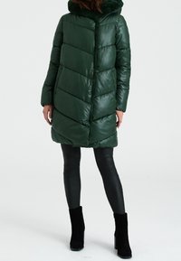 Abrigo de plumas verde con un patrón en forma de chevrón, que presenta una capucha con ribete de piel, cierre de cremallera y mangas largas, combinado con leggins negros ajustados.