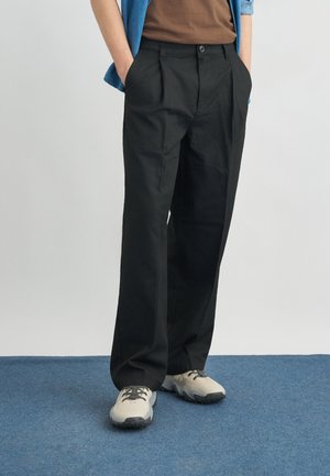 Mężczyzna ubrany w luźne czarne spodnie, beżowo-szare buty sportowe, brązową koszulę oraz niebieską koszulę z długim rękawem, z rękami w kieszeniach, stojący na niebieskim dywanie.