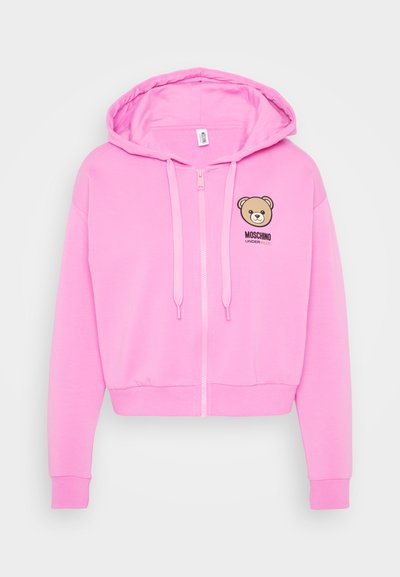 Roza skrajšani dukserica iz mehkega materiala. Ima sprednji zadrgo, kapuco s vrvico in grafiko medvedka s tekstom "Moschino" na prsih.