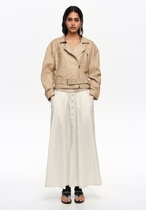 Mujer con chaqueta de cuero beige estilo motorista, falda larga blanca abotonada, calcetines negros y sandalias negras, posando sobre un fondo blanco.