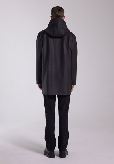 Svart vattentät kappa med huva; har en rak skärning, sömlös design och långa ärmar. Bärs med svarta byxor och stadiga skor.