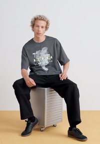 Chemise grise à manches courtes avec un design graphique, accompagnée d'un pantalon noir ample et de baskets noires, assise sur un objet métallique argenté.