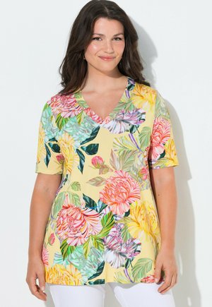 Kvinde iført en gul V-hals kortærmet top med store farverige blomsterprints, kombineret med hvide bukser, stående mod en hvid baggrund.