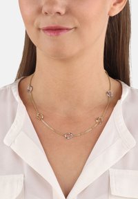 Collar de tono dorado con eslabones redondos alternando entre lisos y retorcidos. El diseño de la cadena presenta acentos plateados. Llevado por una persona con una camisa blanca.