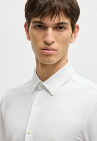 Camicia bianca in tessuto con texture, caratterizzata da colletto classico e bottoni perla, che presenta un design aderente e un tessuto liscio.