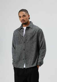 Zwart-wit overhemdjack met visgraatpatroon, een knoopsluiting aan de voorkant, twee borstzakken en een ontspannen pasvorm. De stof heeft een gestructureerde afwerking.