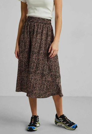 Vrouw staat met een zwarte en roze midi-rok met dierenprint, een wit shirt met korte mouwen en zwart-blauwe sportschoenen.