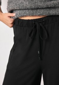 Schwarze Trainingshose mit elastischem Bund, Kordelzugverschluss und seitlichen Taschen. Der Stoff wirkt glatt und hat eine leichte Textur.