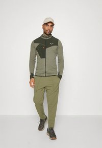 Grønn glidelåsjakke med teksturerte paneler, matchende avslappede olivengrønne bukser og grå joggesko. Antrekket har funksjonelle designelementer.