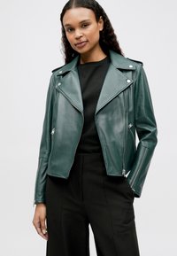 Zelena usnjena motoristična jakna s prilagojeno zasnovo, z zarezanim ovratnikom, žepki z zadrgo in teksturirano površino, nošena čez črno majico.