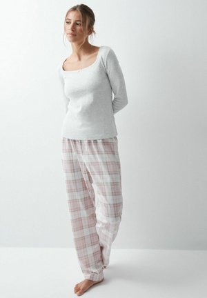 Femme aux cheveux châtain clair portant un haut gris à manches longues et un pantalon de pyjama ample à carreaux roses et blancs, debout pieds nus sur un sol blanc contre un mur blanc.