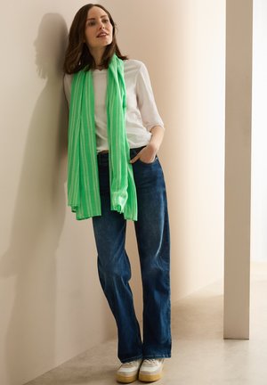Maglietta bianca a maniche lunghe, sciarpa a righe verdi, jeans a gamba larga blu e sneakers bianche con suola beige. Modello appoggiato a un muro chiaro.