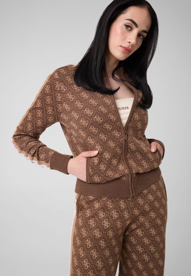 Femme aux cheveux noirs portant une veste zippée marron à motifs et un pantalon assorti, mains dans les poches de la veste, fond gris neutre.