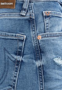 Obszar tylnej kieszeni niebieskich dżinsów z logo True Religion, miedzianymi nitami, pętlą na pasek oraz niewielkim postrzępionym przetarciem po prawej stronie.