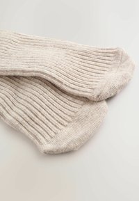 Ein Paar beigefarbene, gerippte Stricksocken flach auf einer weißen Oberfläche ausgelegt, die Textur und Zehendetails zeigen.