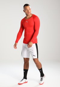 Rød langermet treningstrøye med en tettsittende design, kombinert med hvite shorts med svarte sidestriper, og hvite joggesko med røde detaljer.