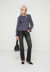 Fiatal nő hosszú szőke hajjal, kék kockás, redőzött felsőt visel, fekete bőrnadrágot, fekete tűsarkú szandált és egy fekete kézitáskát tart.