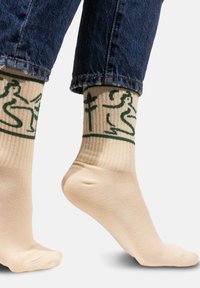 Skarpetki do kolan w kolorze beżowym z zielonymi grafikami przedstawiającymi postacie i rośliny. Żebrowana struktura u góry, wykonane z miękkiego materiału.