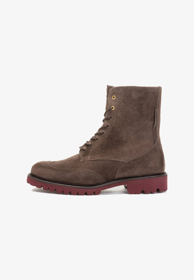 Bottine en suède marron avec un bout arrondi, six œillets en métal et une semelle en caoutchouc rouge robuste. Présente une texture lisse et un design épuré.