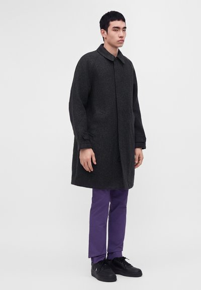Young man wearing a dark gray long coat, purple pants, and black sneakers standing against a plain light background.