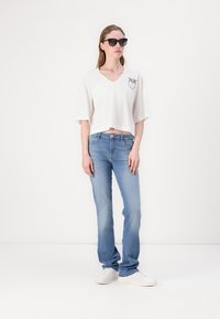 T-shirt bianco corto con scollo a V e grafica di palme, abbinato a jeans blu chiaro, scarpe da ginnastica bianche e occhiali da sole scuri.