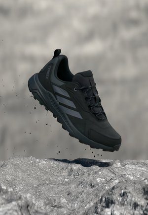 Zapato de senderismo Adidas Terrex negro flotando sobre suelo rocoso con partículas de tierra suspendidas en el aire a su alrededor.