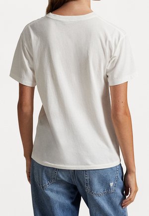 Persoon van achteren gezien die een effen wit T-shirt met korte mouwen draagt en blauwe spijkerbroek met een klein scheurtje op de achterzak.