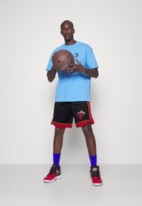 Modrá tričko s logom, čierne šortky s logom Miami Heat a červenými akcentmi, modré ponožky, červeno-čierne basketbalové topánky, drží basketbal.