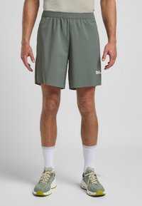 Shorts de sport verts avec une taille élastique, texture lisse et un logo blanc sur la cuisse gauche. Associés à des chaussures de sport gris clair et des chaussettes blanches.