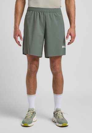 Green athletic shorts with an elastic waistband, smooth texture, and a white logo on the left thigh. Paired with light gray athletic shoes and white socks.