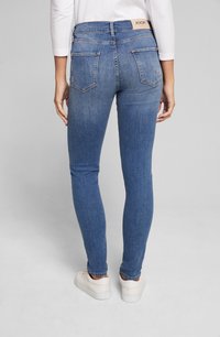 Szczupłe niebieskie dżinsy o gładkiej fakturze, z tylnymi kieszeniami i markową metką na pasku. Noszone z białymi trampkami.