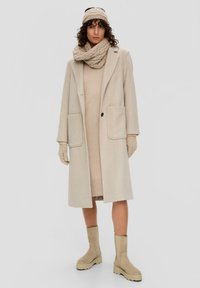 Beige wollen jas met twee voorzakken, gecombineerd met een gebreide trui-jurk, een gestructureerde sjaal en beige laarzen tot de knie.
