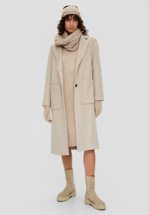 Beige wollen jas met twee voorzakken, gecombineerd met een gebreide trui-jurk, een gestructureerde sjaal en beige laarzen tot de knie.